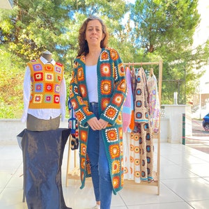 May include: A woman stands in front of a rack of crocheted garments. The garments are made of colorful squares and feature a variety of patterns. The woman is wearing a green crocheted cardigan with a similar square pattern.