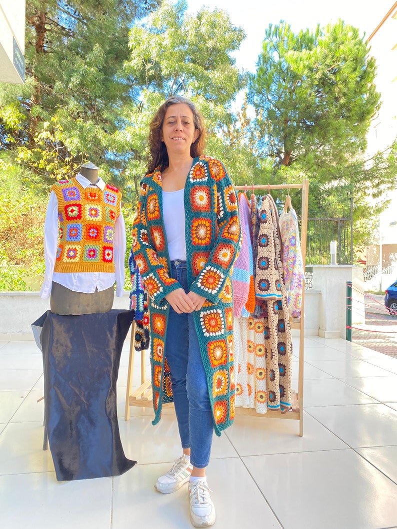 May include: A woman stands in front of a rack of crocheted garments. The garments are made of colorful squares and feature a variety of patterns. The woman is wearing a green crocheted cardigan with a similar square pattern.
