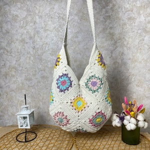 Op de afbeelding: Een witte gehaakte tote bag met een kleurrijk bloemenpatroon. De tas heeft een lange band en is gemaakt van een zacht, lichtgewicht materiaal.