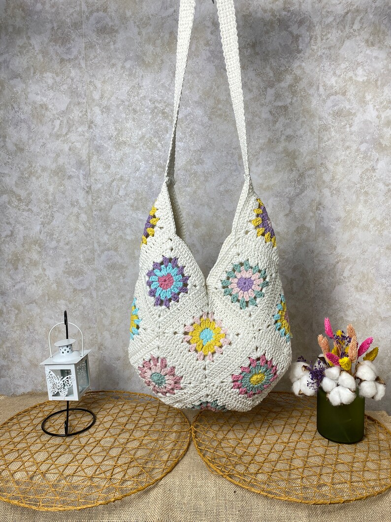 Op de afbeelding: Een witte gehaakte tote bag met een kleurrijk bloemenpatroon. De tas heeft een lange band en is gemaakt van een zacht, lichtgewicht materiaal.