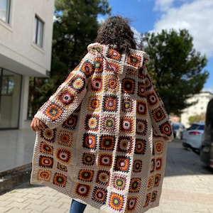 Puede incluir: Un abrigo largo con capucha de ganchillo con un dise&ntilde;o de retazos de cuadrados coloridos en tonos naranja, amarillo, morado y verde. El abrigo es de color crema y parece hecho a mano. Se lleva sobre unos vaqueros azules y zapatillas negras.