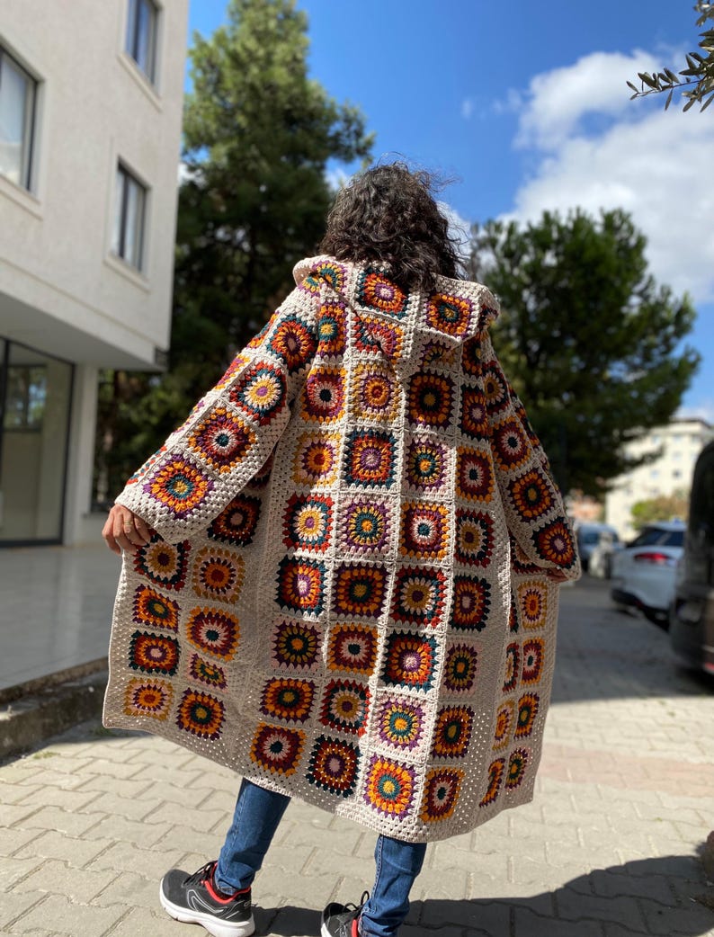 Puede incluir: Un abrigo largo con capucha de ganchillo con un dise&ntilde;o de retazos de cuadrados coloridos en tonos naranja, amarillo, morado y verde. El abrigo es de color crema y parece hecho a mano. Se lleva sobre unos vaqueros azules y zapatillas negras.