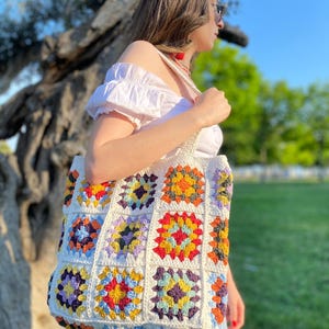 May include: A white crochet tote bag with colorful granny squares. The bag has a large, rectangular shape and is made with a variety of colors, including red, orange, yellow, green, blue, purple, and brown.