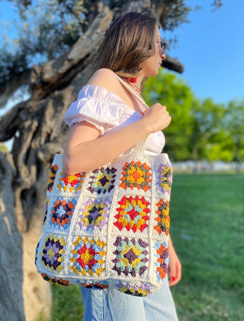May include: A white crochet tote bag with colorful granny squares. The bag has a large, rectangular shape and is made with a variety of colors, including red, orange, yellow, green, blue, purple, and brown.