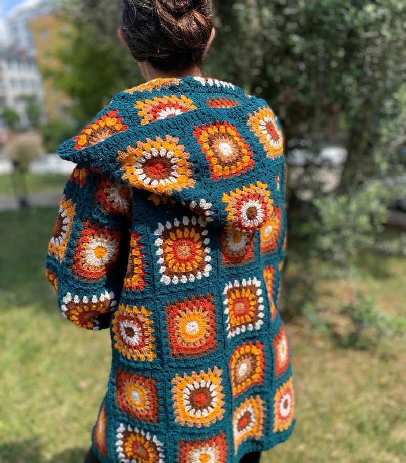 K&ouml;nnte beinhalten: Eine tealblaue geh&auml;kelte Strickjacke mit Kapuze. Die Strickjacke ist mit einem Granny-Square-Muster gearbeitet und hat orange, braune und wei&szlig;e Akzente.