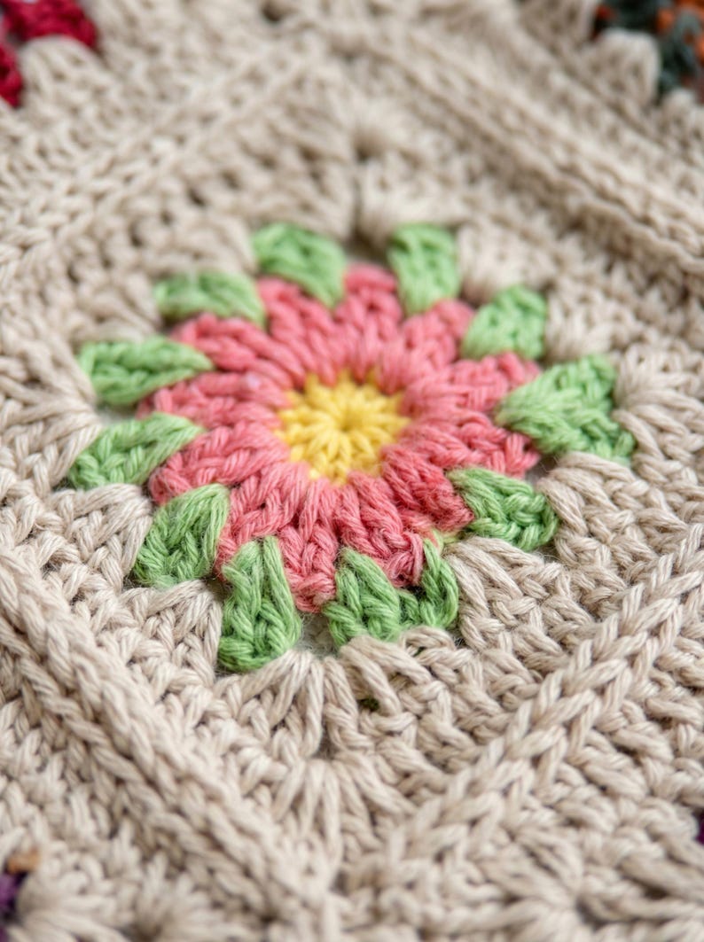 May include: Close-up of a crocheted square featuring a floral design. The flower has a yellow centre, pink petals, and green leaves. The surrounding crochet is a neutral beige colour, creating a textured, handmade appearance. Ideal for home decor.