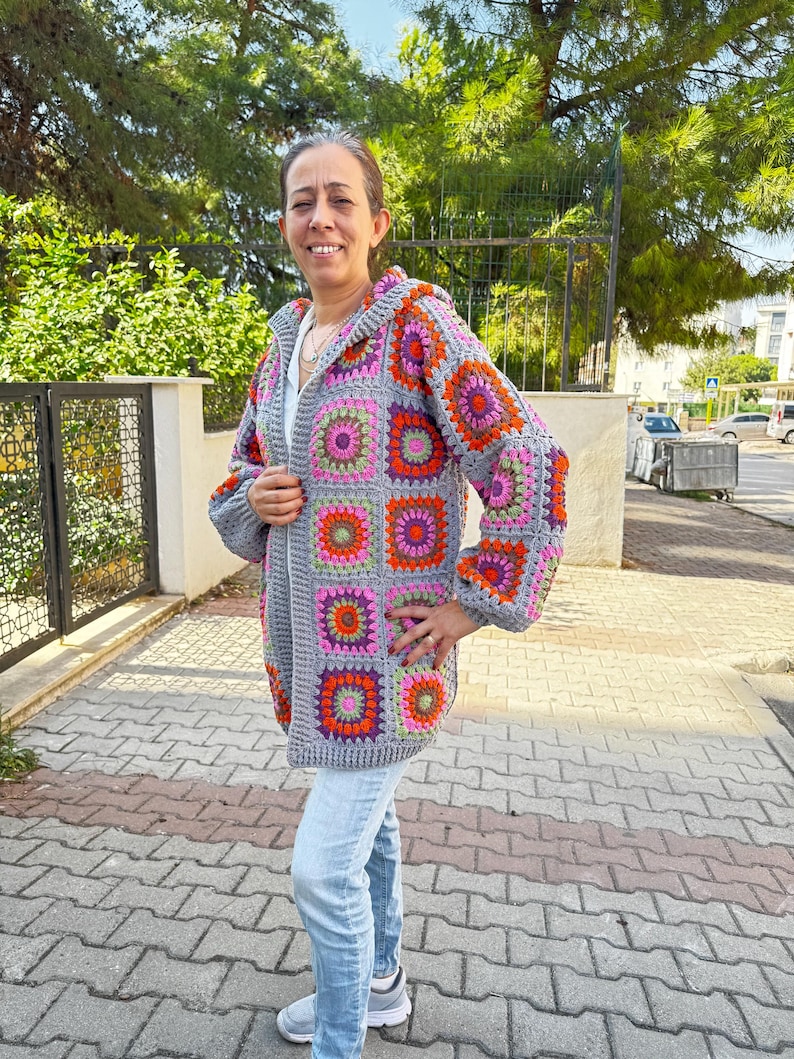 May include: A gray crocheted cardigan with a hood, featuring a colorful geometric pattern of squares in shades of pink, orange, green, and purple.