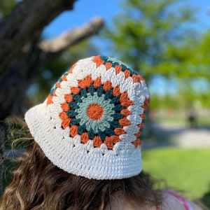 Puede incluir: Un sombrero de cubo de crochet blanco con un patr&oacute;n geom&eacute;trico colorido en tonos naranja, verde y blanco.