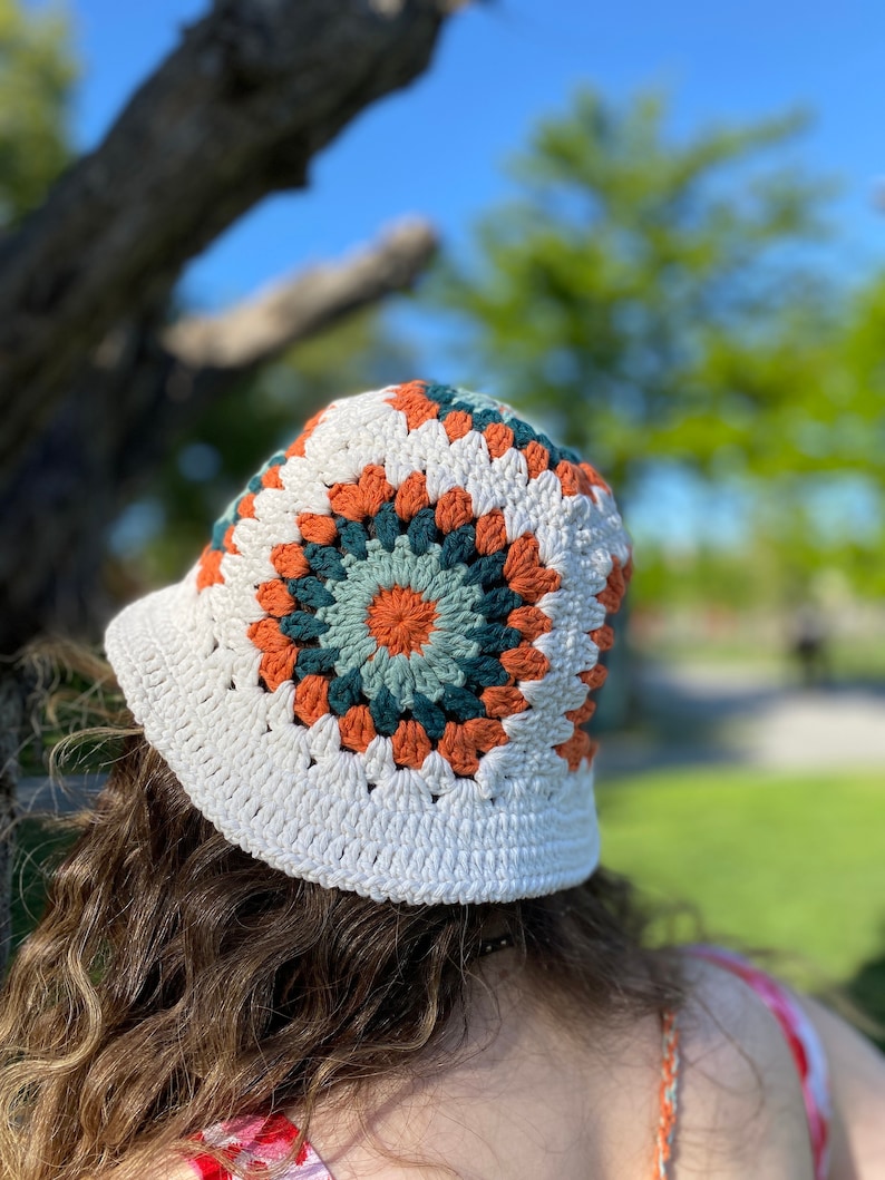 Puede incluir: Un sombrero de cubo de crochet blanco con un patr&oacute;n geom&eacute;trico colorido en tonos naranja, verde y blanco.