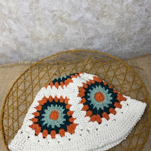 Puede incluir: Sombrero blanco de crochet en forma de cubo con un patr&oacute;n geom&eacute;trico en tonos naranja, verde y marr&oacute;n.