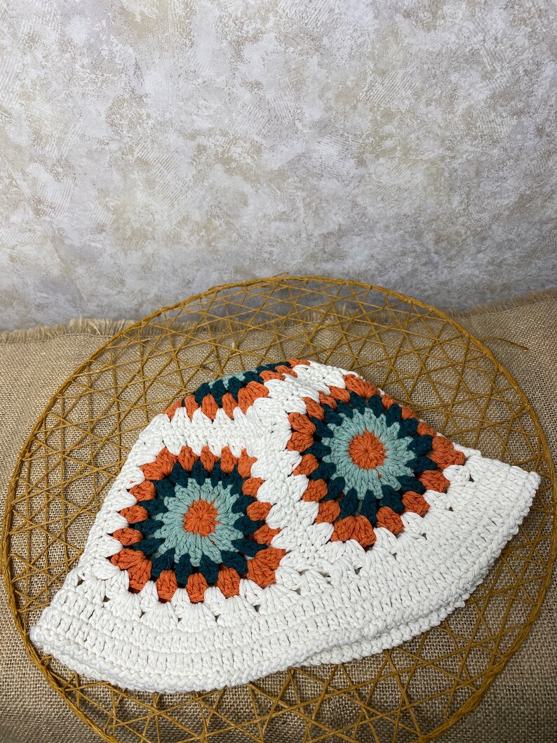 Puede incluir: Sombrero blanco de crochet en forma de cubo con un patr&oacute;n geom&eacute;trico en tonos naranja, verde y marr&oacute;n.