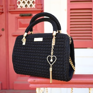 May include: A black crochet handbag with gold-colored hardware, including a chain strap and a heart-shaped charm with a pearl and tassel. The bag has two braided handles and a rectangular shape.