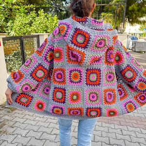 May include: A gray crochet cardigan with a hood, featuring a colorful geometric pattern of squares with pink, orange, green, and purple accents.