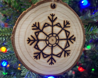 Op de afbeelding: Een houten kerstversiering met een sneeuwvlokdesign dat in het oppervlak is gebrand. De versiering hangt aan een kerstboom met kleurrijke lampjes.