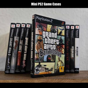 Puede incluir: Una colección de mini estuches de juegos de PS2, incluyendo "Grand Theft Auto: San Andreas" y otros. Los juegos se muestran en una estantería de madera contra una pared blanca. El estuche de "Grand Theft Auto" es el más prominente, con una portada colorida.