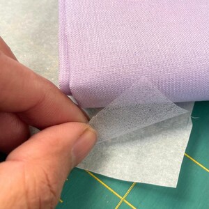 May include: A hand peeling a thin, translucent layer from a folded piece of light purple fabric. The fabric is a solid colour, possibly cotton or linen. The background is a light grey surface, possibly a cutting mat.