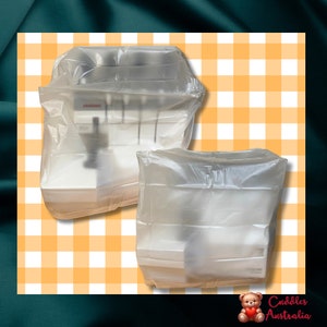 May include: Two white sewing machines in clear plastic packaging on a yellow and white checkered background. The machines are both wrapped in clear plastic and have the word "Janome" printed on them.