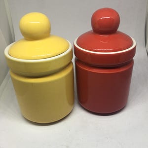 May include: Two ceramic canisters with lids. The canister on the left is yellow and the canister on the right is red.