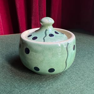 May include: A round, lidded ceramic jar with a light green glaze. The jar is decorated with dark blue polka dots and a wavy grey line. The lid has a small knob and matching polka dots.