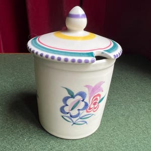 May include: A white ceramic jar with a lid, decorated with a floral pattern in blue, pink, and purple. The lid has a colourful rim with a purple and teal dotted border. The jar has a small spoon rest.