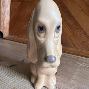 Könnte beinhalten: Eine Keramik-Hundefigur in sitzender Position. Der Hund hat ein hellbraunes Fell, lange Schlappohren und blaue Augen. Die Nase ist dunkler. Die Figur steht auf einer Holzoberfläche.