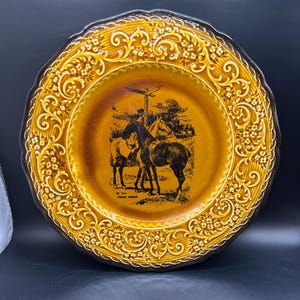 May include: A decorative plate with a brown and gold color scheme. The plate features a central illustration of a person on horseback, with the text "ST. THE CROSS ROADS" below the image. The plate has a scalloped edge and ornate floral detailing.