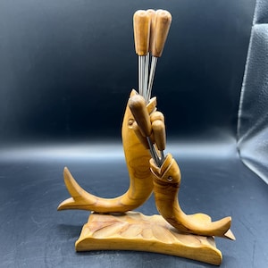 May include: A wooden fondue set featuring two stylized fish holding fondue forks. The fish are carved from light-colored wood and rest on a rectangular base with leaf detailing. The forks have wooden handles and stainless steel tines.