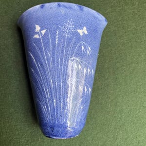 May include: A blue ceramic wall pocket with a textured finish. The vase features a white floral and butterfly design. The pocket is shaped like a vase and is designed to be hung on a wall. The overall theme is nature and home decor.