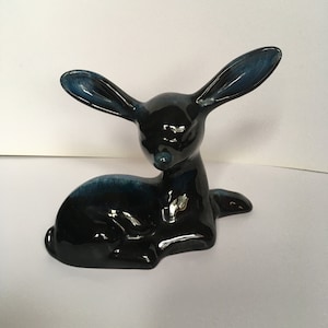 May include: A ceramic deer figurine in a reclining pose. The deer is primarily black with glossy, dark blue accents on the ears and back. The figurine has a smooth, reflective surface and is set against a plain white background.