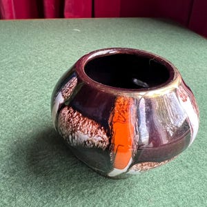 May include: A small, round, ceramic planter with a dark brown glaze and abstract geometric patterns in white, orange, and brown.