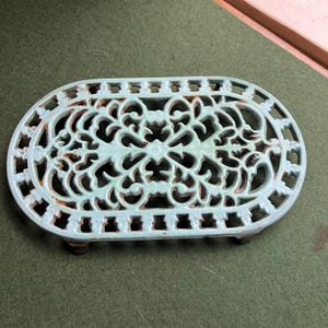 May include: A light blue, oval trivet with an intricate, openwork pattern. The metal trivet has a distressed finish, showing signs of rust. It is supported by small feet. The trivet is designed to protect surfaces from heat.