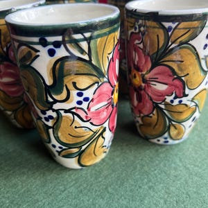 May include: A collection of six ceramic tumblers. Each tumbler has a white base adorned with a floral pattern in pink, yellow, and green. The rims are green, and the design includes blue dots. The tumblers are on a green surface.