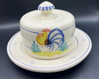 May include: White ceramic cheese dome with a rooster design. The dome has a blue and white trim and a rooster with a blue tail and yellow body. The dome sits on a matching plate.