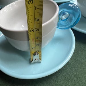 May include: White and light gray teacups with blue saucers. The teacup is approximately 2 inches (5 cm) tall. The handle is a transparent blue. The teacups are on a green surface.
