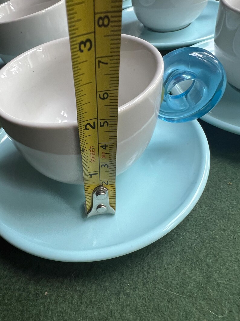 May include: White and light gray teacups with blue saucers. The teacup is approximately 2 inches (5 cm) tall. The handle is a transparent blue. The teacups are on a green surface.