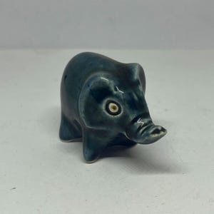 May include: A small, ceramic elephant figurine in a dark teal color. The elephant has a rounded body, small legs, and a trunk. The eyes are detailed with a white center and a dark outer ring. The figurine is glossy and appears to be handmade.