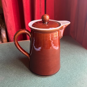 May include: A brown ceramic pitcher with a lid and handle. The pitcher has a glossy finish and a white rim. The lid is topped with a small, round knob. The pitcher is designed for pouring liquids.