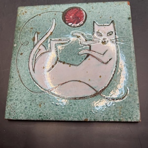 May include: Square ceramic tile with a playful cat design. The cat is white with a light blue outline, playing with a red ball of yarn. The background is a speckled teal color. A stylized cat illustration.