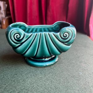 May include: A teal ceramic bowl with a glossy finish, featuring a shell-like design with vertical fluting and prominent spiral scrolls on each side. The bowl is supported by a circular pedestal base and has a wide, open top with a scalloped rim.