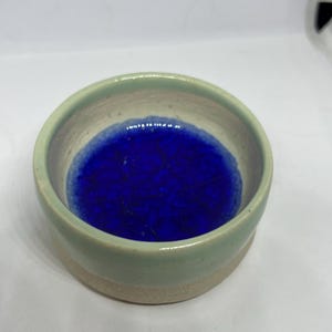 May include: A small, handmade ceramic bowl. The exterior is light green, and the interior is a vibrant blue. The bowl has a simple, rounded shape and a natural, unglazed base. The blue glaze has a textured, crackled appearance.
