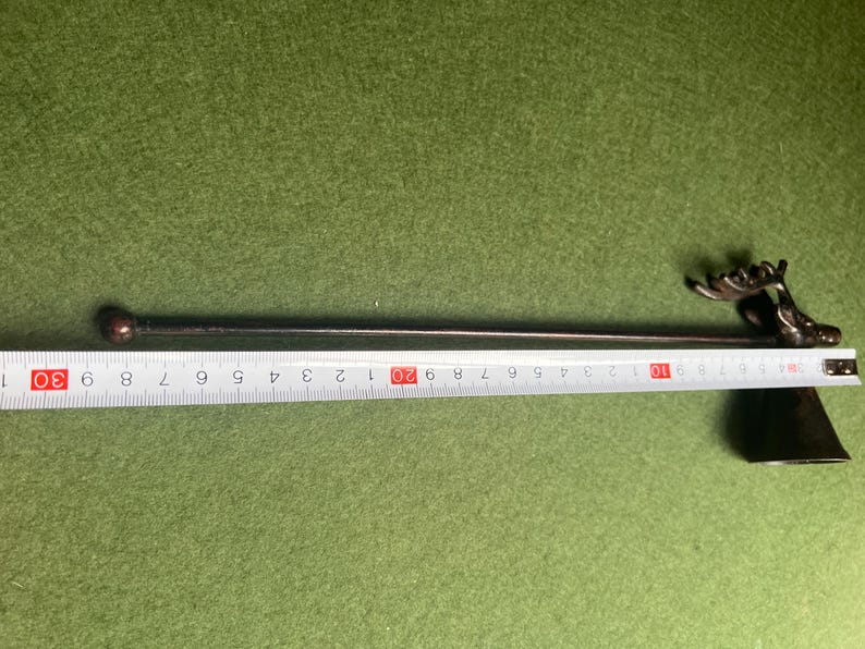 Może przedstawiać: Metalowy pogrzebacz do kominka z dekoracyjnym motywem głowy łosia. Pogrzebacz ma około 74 cm długości, z okrągłą kulką na przeciwległym końcu głowy łosia. Pogrzebacz leży na zielonej powierzchni.