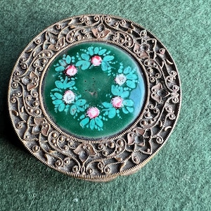 Puede incluir: Un compacto redondo y antiguo con un marco de bronce ornamentado y detallado. El centro presenta un fondo verde con un diseño floral de flores rosas y blancas. El compacto está sobre un fondo verde.