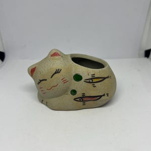 May include: A beige ceramic cat-shaped planter with a sleeping expression. The planter features painted details including red ears, a smiling face, and fish designs. The planter has a round opening for plants.