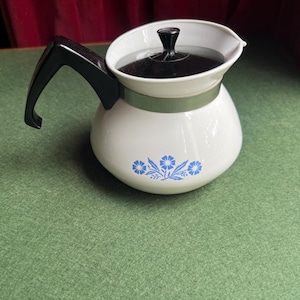 May include: A white teapot with a rounded body featuring a blue floral pattern. It has a black handle, a black lid with a central knob, and a silver-colored metal band encircling the upper portion of the pot below the lid opening.