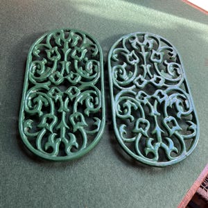 May include: Two green cast iron trivets with an ornate, openwork floral design. Each oval-shaped trivet features a detailed pattern of swirling leaves and flowers. The trivets are displayed on a green surface.