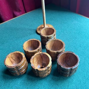 May include: Six small, hand-carved wooden cups with a barrel-like design. The cups are arranged on a green surface, with a wooden stand and a long wooden stick in the background. The wood has a warm, natural tone.