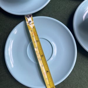 May include: A light blue saucer with a diameter of approximately 5 inches. The saucer is part of a set, with other saucers and a teacup visible. A yellow measuring tape is placed across the saucer.