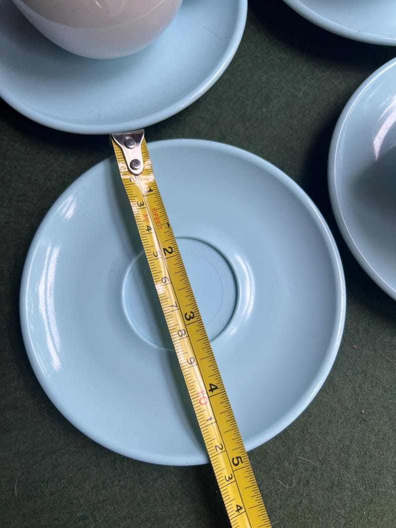 May include: A light blue saucer with a diameter of approximately 5 inches. The saucer is part of a set, with other saucers and a teacup visible. A yellow measuring tape is placed across the saucer.