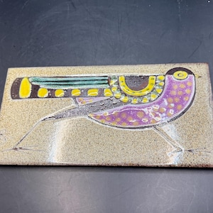 May include: A rectangular ceramic tile with a bird design. The bird has a purple body with yellow spots, a yellow and black head, and a yellow and green wing. The tile has a beige background and black legs.