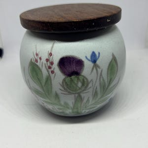May include: A small, round ceramic jar with a wooden lid. The jar is decorated with hand-painted floral designs, including purple thistles, blue flowers, and green leaves. The lid is a dark brown color.